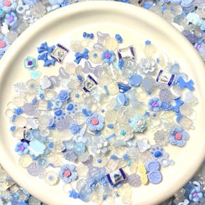 May include: A white bowl filled with a variety of blue and white resin embellishments. The embellishments include flowers, bows, stars, and other shapes. The bowl is surrounded by more embellishments.