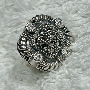 May include: A silver ring with a square-shaped setting featuring a black marcasite stone surrounded by four smaller white stones.