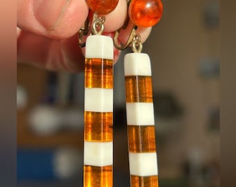 May include: A pair of clip-on earrings with amber and white striped rectangular pendants. The amber is a deep orange color and the white stripes are opaque.
