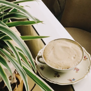 May include: A close-up of a cup of coffee with a light brown frothy top, sitting on a white saucer with a floral pattern. The saucer is on a white table with a green plant in the background.