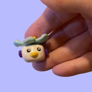 May include: A small, handmade figurine of a cartoon character with a white body, yellow beak, and purple ear-like details. The character has a light blue headpiece and is held in a hand.