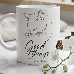 Könnte beinhalten: Ein weißer Keramikbecher mit einer schwarzen Strichzeichnung einer Katze, die einen Schmetterling betrachtet. Der Text Good things take time ist in schwarzer Schreibschrift unter der Katze gedruckt. Der Becher steht neben einer weißen Orchidee.