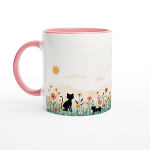 May include: A white ceramic mug with a pink handle and interior rim. The exterior features a printed illustration of two black cat silhouettes sitting in a field of colorful wildflowers under a yellow sun and pale clouds against a light background.