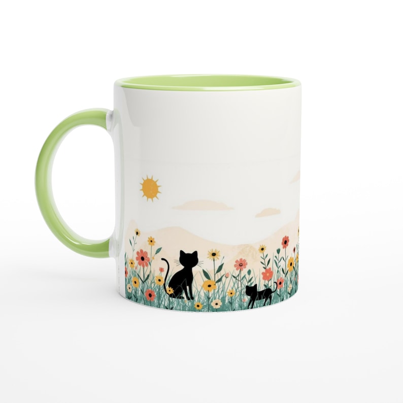 May include: A white ceramic mug with a light green handle and interior rim. The exterior features a printed illustration of two black cats sitting in a field of colorful wildflowers under a yellow sun and pale clouds against a soft beige background.