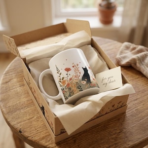 May include: A white ceramic mug featuring a black cat illustration surrounded by orange and red wildflowers, nestled in a brown cardboard gift box with white tissue paper and a small card printed with the text for you my dear.