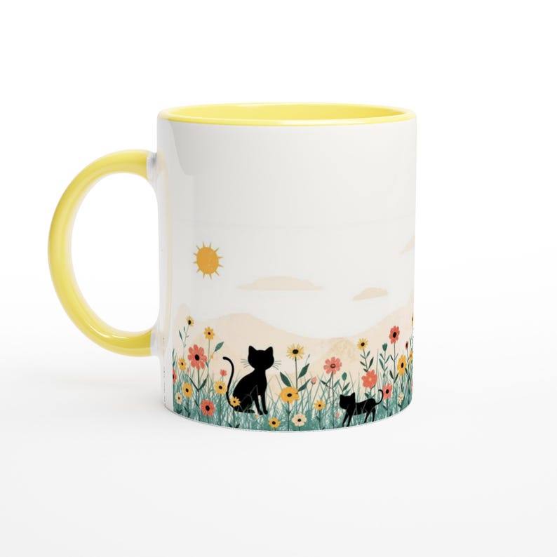 May include: A white ceramic mug with a bright yellow handle and interior rim. The exterior features a colorful illustration of two black cats sitting in a field of wildflowers under a yellow sun, set against a soft, pale landscape background.