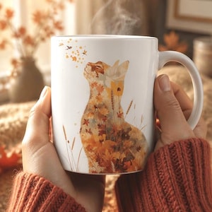 Könnte beinhalten: Ein weißer Keramikbecher mit einer grafischen Illustration einer Katze, die aus Herbstblättern und Weizenhalmen besteht. Die Katze blickt nach oben zu einer kleinen Ansammlung schwebender Blätter. Der Becher hat einen geschwungenen Henkel.