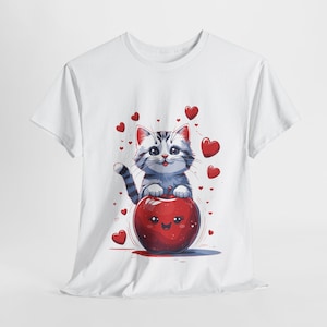 Könnte beinhalten: Weißes T-Shirt mit einem Cartoon-Bild eines grau-weißen Katers, der auf einem roten Apfel mit einem lächelnden Gesicht sitzt. Der Kater ist von kleinen roten Herzen umgeben.