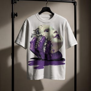 Puede incluir: Camiseta blanca con un gráfico surrealista de una cara y efectos de pintura morada. El diseño incluye una cara con rasgos agrietados y una mano que emerge de la pintura. La camiseta está colgada en una percha negra.