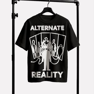 Peut inclure: T-shirt noir avec les mots "ALTERNATE REALITY" en blanc. Un graphisme blanc d'une personne couronnée et d'autres figures est centré sous le mot "ALTERNATE". Le t-shirt est suspendu à un cintre noir.
