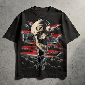Puede incluir: Camiseta negra con un diseño gráfico de una calavera con ojos grandes y boca abierta, superpuesta a una cruz envuelta en alambre de púas. El fondo presenta nubes rojas y negras, creando una estética oscura y ominosa.