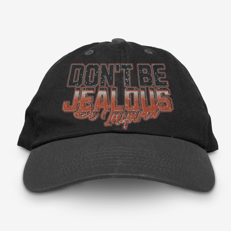 May include: Black baseball cap with the text "Don't Be Jealous Be Inspired" in orange and white lettering.