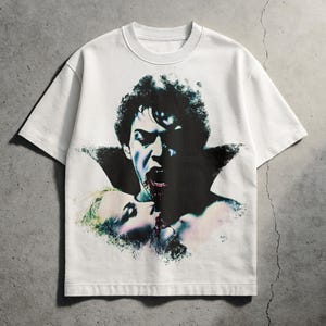 May include: White t-shirt featuring a graphic of a vampire biting a person. The vampire has dark hair and a black cape. The other person has blonde hair. The image has a vintage, horror movie aesthetic.