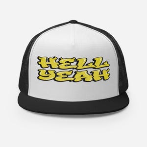 May include: A black and white trucker cap featuring a white front panel with the text HELL YEAH printed in a bold yellow graffiti-style font with black outlines. The cap includes a black mesh back, a black brim, and a structured five-panel design.