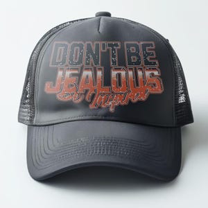 May include: A black trucker hat with a mesh back. The front of the hat has the text "Don't Be Jealous Be Inspired" in white and orange lettering.