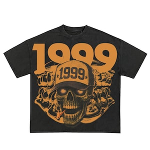 Puede incluir: Camiseta negra con un gráfico desgastado que presenta una calavera con una gorra de béisbol con "1999" en ella. El número "1999" también está impreso en letras grandes de color naranja encima de la calavera. La calavera está fumando un cigarrillo.