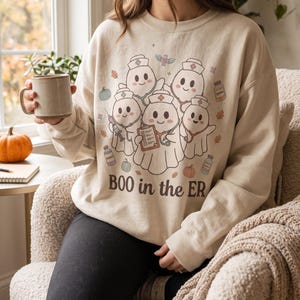 Puede incluir: Una sudadera beige de cuello redondo con un gráfico de cinco fantasmas de dibujos animados vestidos de enfermeros con estetoscopios, una jeringuilla y un portapapeles. El texto "BOO in the ER" está impreso bajo los fantasmas, rodeado de calabazas.