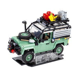 May include: A green LEGO Land Rover Defender 90 model with a black roof rack and accessories. The model is on a black display stand with a clear acrylic plaque that reads "LAND ROVER CLASSIC DEFENDER 90 10317".