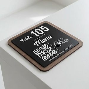 Op de afbeelding: Een vierkant tafelbordje met een donkerhouten voet en een zwart bovenpaneel. Het bordje bevat de witte tekst Table 105, het woord Menu, een QR-code, een NFC-pictogram en de Franse tekst Scannez le QR code ou utilisez le NFC in het wit.