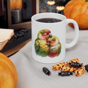 Könnte beinhalten: Eine weiße Keramiktasse mit einer bunten Illustration von zwei kuschelnden Papageien. Die Papageien sind grün, gelb, rot und orange. Die Tasse hat einen schwarzen Henkel und ist mit Kaffee gefüllt.