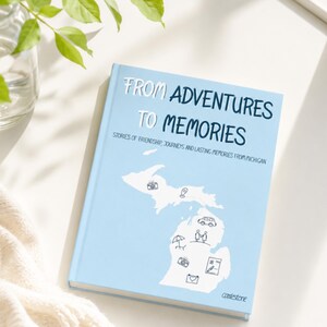 Könnte beinhalten: Ein hellblaues Hardcover-Buch mit dem Titel FROM ADVENTURES TO MEMORIES, STORIES OF FRIENDSHIP, JOURNEYS AND LASTING MEMORIES FROM MICHIGAN. Das Cover zeigt eine weiße Silhouette des Bundesstaates Michigan mit kleinen Strichzeichnungen von Symbolen.