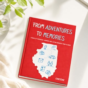 Könnte beinhalten: Ein rotes Hardcover-Buch mit dem Titel FROM ADVENTURES TO MEMORIES, STORIES OF FRIENDSHIP, JOURNEYS AND LASTING MEMORIES FROM ILLINOIS. Das Cover zeigt einen weißen Umriss des Bundesstaates Illinois mit blauen Liniensymbolen einer Landkartenmarkierung, Kamera, Auto, Umschlag, Personen und Stift.