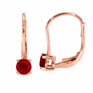 May include: A pair of rose gold leverback earrings, each featuring a single round-cut, deep red gemstone held in a four-prong setting. The metal has a polished, reflective finish, and the design includes a curved wire mechanism for secure closure.