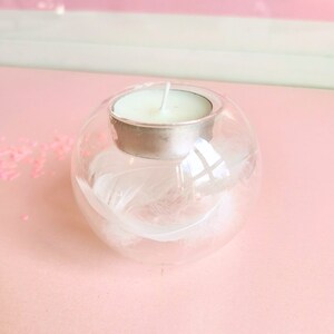 May include: A clear glass candle holder with a white feather inside. The holder has a silver metal top and a white tea light candle inside.
