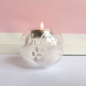 May include: A clear glass candle holder with a white paw print and the name "Bella" etched on the side. The holder is filled with white feathers and a small tea light candle is burning inside.