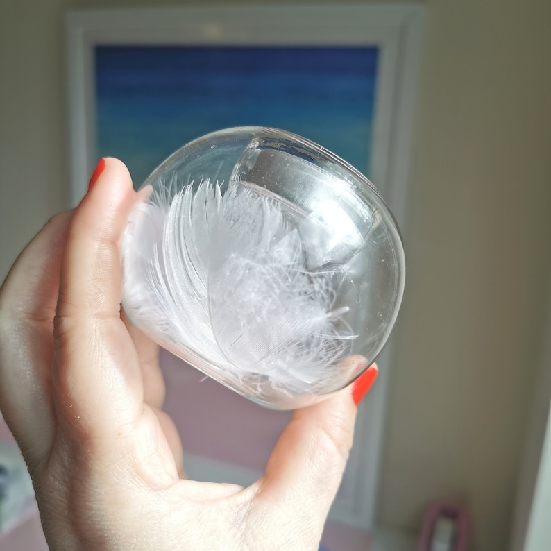 May include: A clear glass jar with a white feather inside. The jar is being held by a hand with red nail polish.