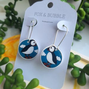 May include: A pair of silver dangle earrings with blue and white puffins on round, white, resin charms. The earrings are displayed on a white card with the text "FOX & BUBBLE Handmade Crafts".