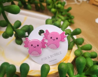 May include: A pair of pink axolotl stud earrings. Each earring features a cartoon axolotl design with a smiling face and frilly gills. The earrings are displayed on a white card with the text "FOX & BUBBLE Handmade Crafts".