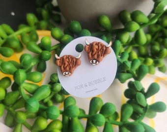 May include: A pair of brown and tan Highland cow earrings. The earrings are on a white card with the text "FOX & BUBBLE". The earrings are surrounded by green foliage.