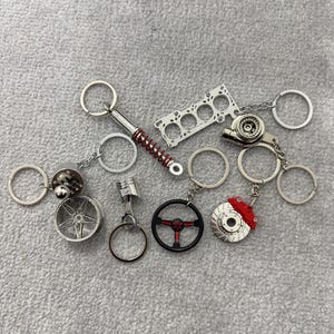 Pode incluir: Uma coleção de chaveiros de metal prateado com peças de carro em miniatura. Os chaveiros incluem uma roda, um pistão, um volante, um disco de freio com pinça vermelha e um turbocompressor. Outros chaveiros incluem uma mola e uma cabeça de cilindro.