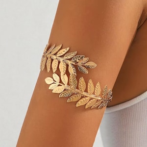May include: Gold-colored arm cuff featuring a leaf design. The cuff wraps around the upper arm, with detailed leaf patterns. The jewelry is made of a shiny, metallic material, and is worn on a tanned arm.