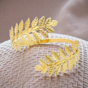 May include: Gold-colored leaf-shaped cuff bracelet. The open-ended bracelet features detailed leaf designs on each end, connected by a smooth, curved band. The bracelet is displayed on a textured, beige surface.