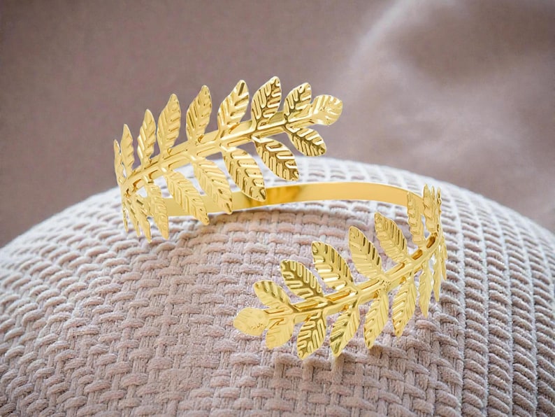 May include: Gold-colored leaf-shaped cuff bracelet. The open-ended bracelet features detailed leaf designs on each end, connected by a smooth, curved band. The bracelet is displayed on a textured, beige surface.