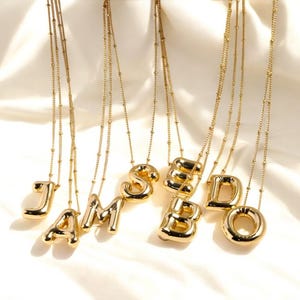 Puede incluir: Un conjunto de seis collares de tono dorado con colgantes de letras. Las letras deletrean la palabra "JAMBOD".