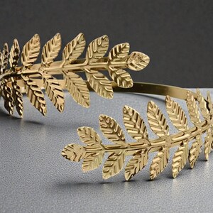 May include: A gold-colored arm cuff featuring a laurel leaf design. The cuff is made of metal and has a flexible band. The leaves are detailed with veins and are arranged in a symmetrical pattern. The cuff is designed to be worn on the upper arm.