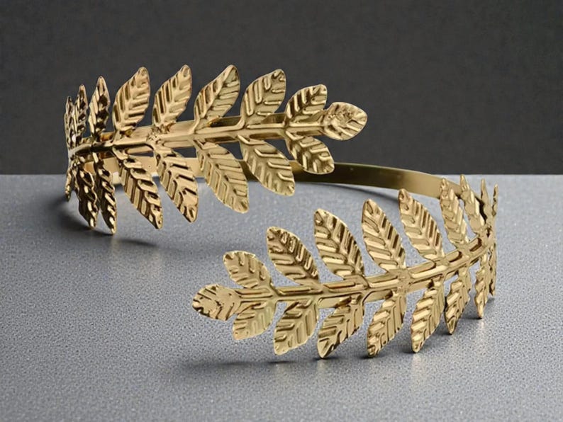 May include: A gold-colored arm cuff featuring a laurel leaf design. The cuff is made of metal and has a flexible band. The leaves are detailed with veins and are arranged in a symmetrical pattern. The cuff is designed to be worn on the upper arm.