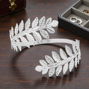 May include: A silver-colored arm cuff featuring a laurel leaf design. The cuff is open, with leaf clusters at each end. The jewelry is displayed on a dark wooden surface, with a jewelry box in the background.
