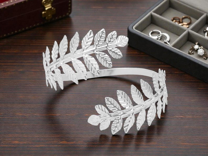 May include: A silver-colored arm cuff featuring a laurel leaf design. The cuff is open, with leaf clusters at each end. The jewelry is displayed on a dark wooden surface, with a jewelry box in the background.