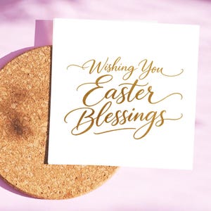 May include: A white card with gold script reading "Wishing You Easter Blessings" sits on a round cork coaster. The background is a soft pink, with shadows adding depth. A seasonal greeting for Easter.