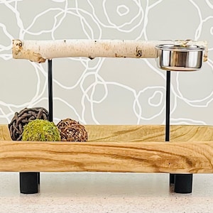 May include: A rectangular wooden bowl with a natural grain finish, supported by four black cylindrical feet. A horizontal birch branch is mounted on two black metal rods, featuring a small silver metal cup attached to the right side.