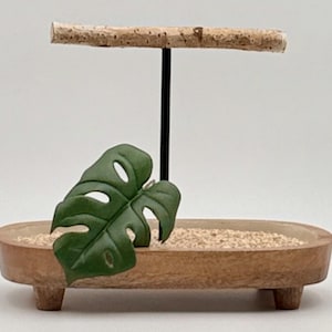 May include: An oval wooden tray with four short feet, filled with light-colored sand. A black metal rod stands vertically in the center, topped with a horizontal piece of natural bark-covered wood. A green monstera leaf rests inside.