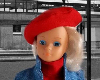 May include: A doll wearing a red beret, a red turtleneck, and a denim jacket. The doll has blonde hair and blue eye shadow. The background is a black and white street scene.