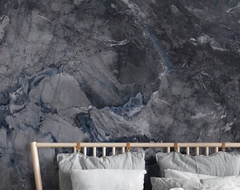 May include: A bedroom scene with a bed featuring grey bedding and a wooden headboard. The wall behind the bed has a dark grey marble design with blue veining. A large green plant is in the corner.