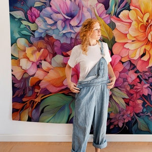 May include: A woman wearing light blue denim overalls and a white t-shirt stands in front of a large floral tapestry. The tapestry features a watercolor design with pink, orange, and purple flowers and green leaves.