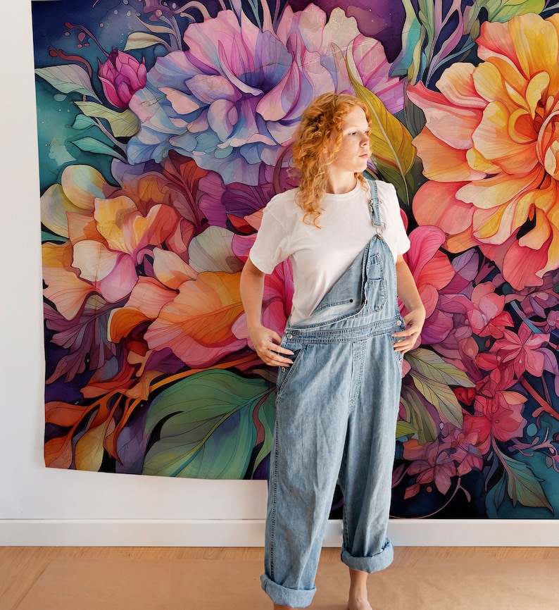 May include: A woman wearing light blue denim overalls and a white t-shirt stands in front of a large floral tapestry. The tapestry features a watercolor design with pink, orange, and purple flowers and green leaves.