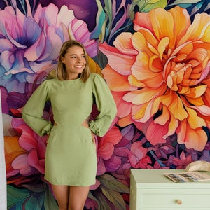 May include: A woman in a light green dress stands in front of a floral mural. The mural features large, colorful flowers in shades of pink, purple, and orange. A light green cabinet with a few items on top is visible.